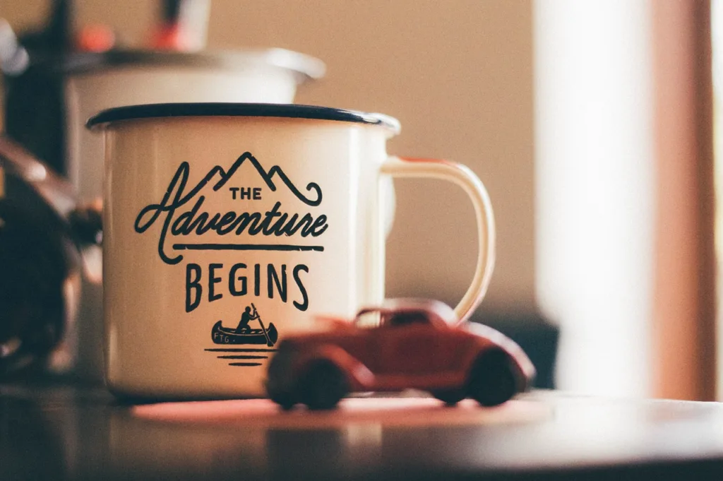 White enamel mug with 'The Adventure Begins' printed on it, sitting on a table with a small red toy car, symbolizing a new journey and inspiring a happy journey quote.