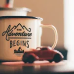 White enamel mug with 'The Adventure Begins' printed on it, sitting on a table with a small red toy car, symbolizing a new journey and inspiring a happy journey quote.