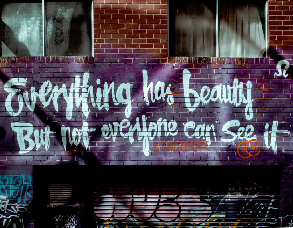 Street art graffiti on a brick wall reading 'Everything has beauty, but not everyone can see it,' with artistic purple and white lettering.
